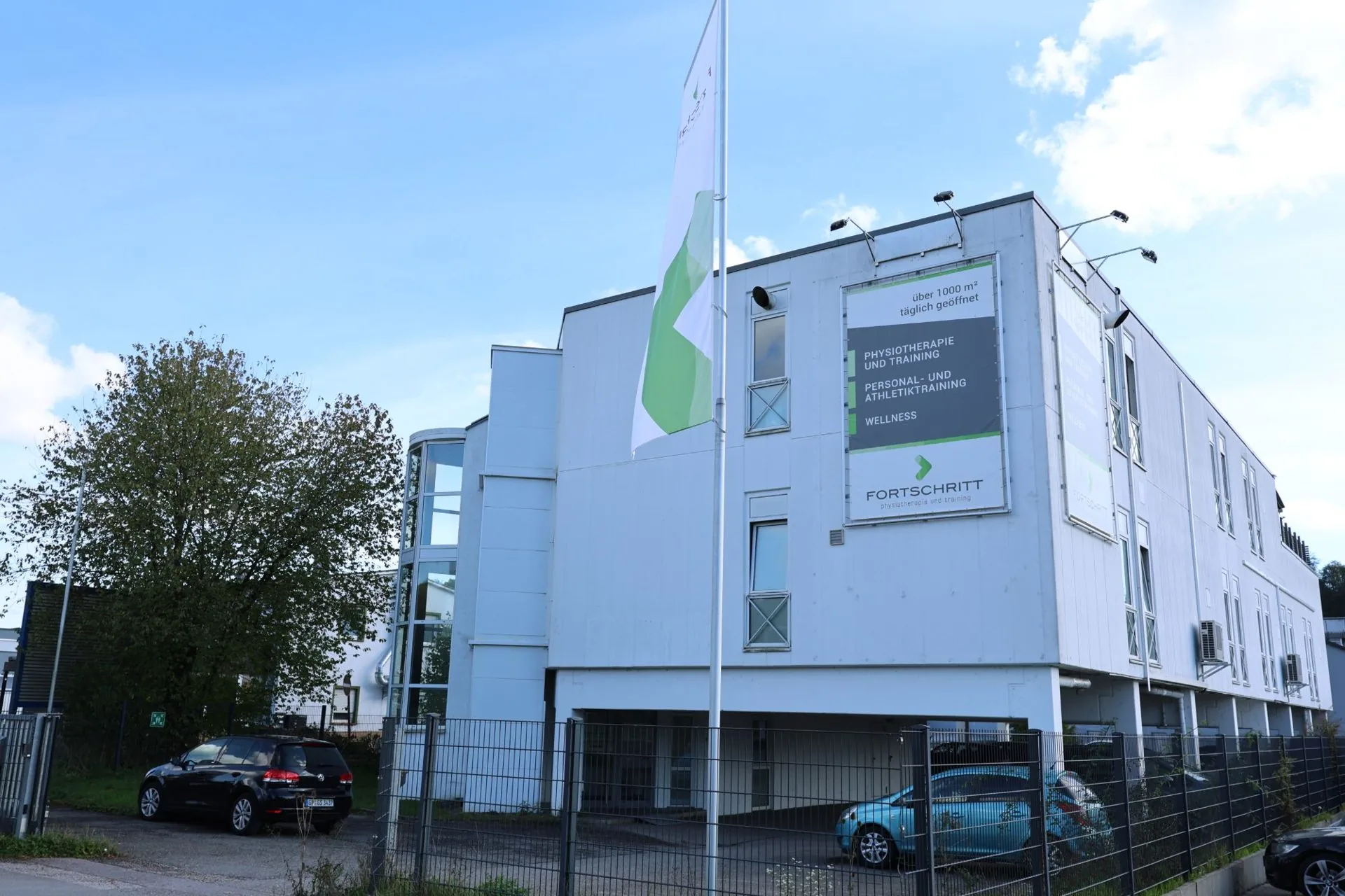 Außenansicht FORTSCHRITT Ebersbach: weißes Gebäude mit grünem FORTSCHRITT-Banner, Fahne und Parkplätzen