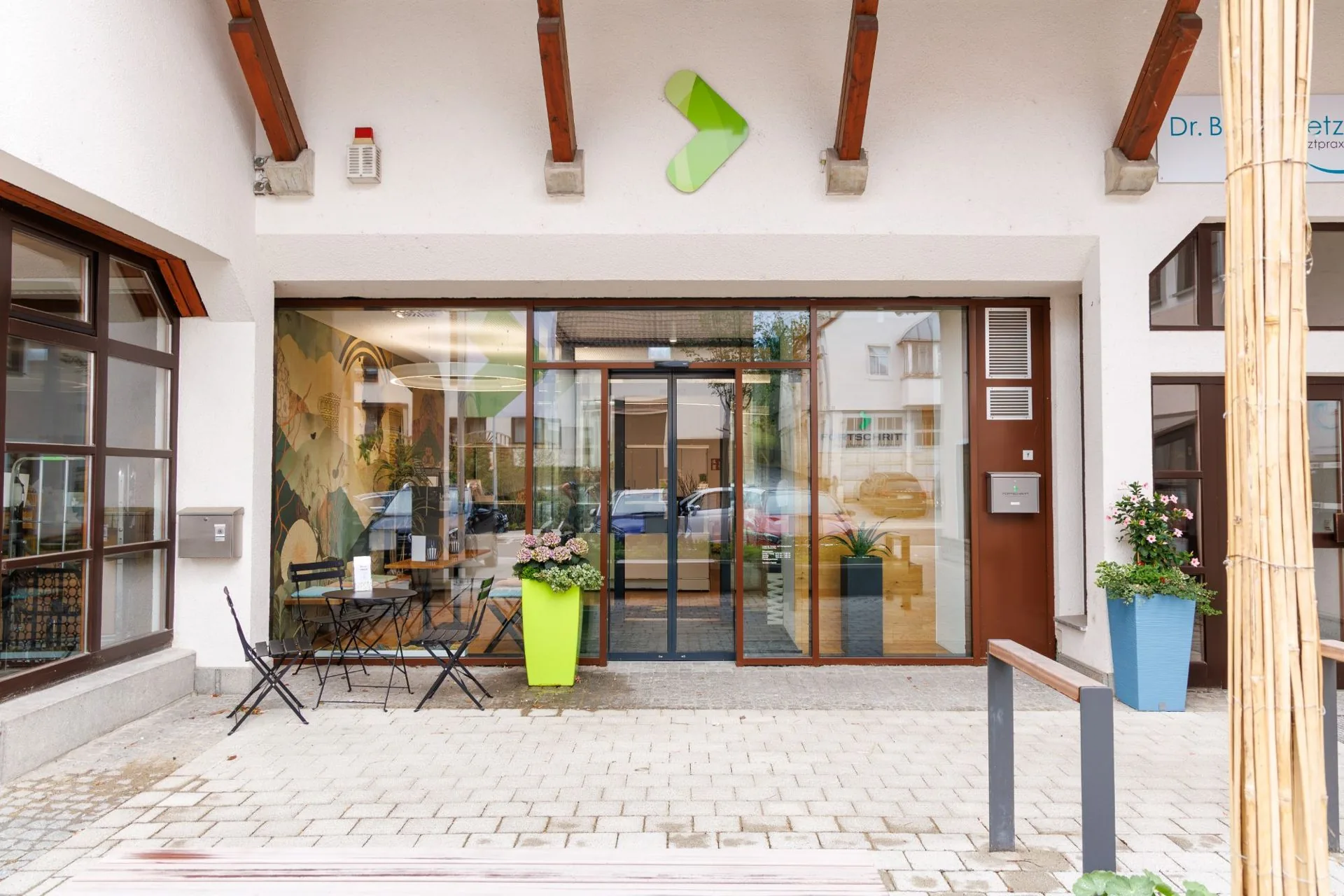 Eingang FORTSCHRITT Schlierbach: Glastür mit grünem FORTSCHRITT-Pfeil, Bistrotisch und Blumenkübeln